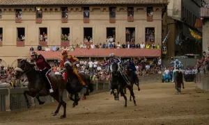 Cavalli e fantini in gara durante le contrade di Siena, con spettatori affacciati ai palazzi.