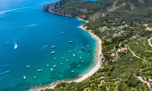 Baia con acque cristalline e numerose barche al largo della costa del Monte Argentario. Ville immerse nel verde e una strada panoramica.