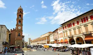 Piazza gremita a Faenza, Italia, con un mercato vivace sotto un cielo azzurro. La torre dell'orologio domina la scena.
