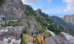 Bicicletta gialla in primo piano, con un panorama mozzafiato di montagne scoscese e un passo del vestito che si snoda tra le rocce.