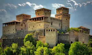 Rocche imponenti e torri medievali, tra i borghi vicino Parma, si stagliano contro un cielo nuvoloso, circondate da un verde lussureggiante.