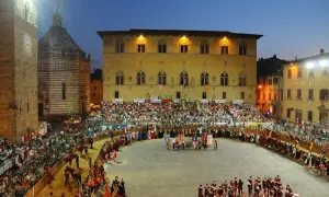 La giostra dell'orso in piazza, con figuranti in costume, cavalli e tribune gremite di spettatori sotto il cielo serale.
