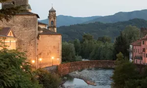 Ponte in pietra sul fiume, con edifici storici illuminati e il monte Argenia sullo sfondo.