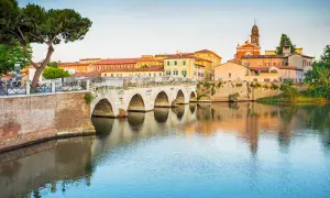 Il Ponte di Tiberio a Rimini, con i suoi archi in pietra che si specchiano sull'acqua calma, fa da sfondo a un paesaggio urbano pittoresco.