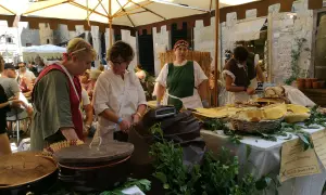 Donne in costume d'epoca servono cibo a un **Volterra markt**. Cesti, pentole e verdure fresche decorano il banco.