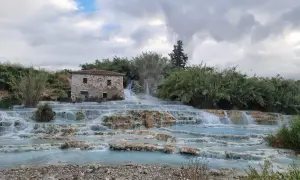 Cascate naturali in Toscana con un antico casale in pietra. L'acqua azzurra scorre su rocce calcaree, creando piscine termali dove alcune persone si rilassano.