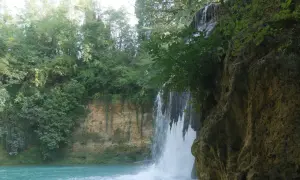 Cascata nel parco fluviale dell'Elsa, con acqua turchese e vegetazione lussureggiante che incornicia la roccia.