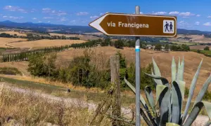 Segnale marrone indica la via Francigena, con un paesaggio collinare toscano sullo sfondo.