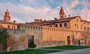 Antica cinta muraria e torri in mattoni rossi sotto un cielo azzurro striato di nuvole rosa, tipico delle colline senesi al tramonto.