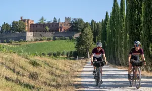Due ciclisti affrontano un eroico percorso su una strada sterrata in Toscana, con un castello e vigneti sullo sfondo.