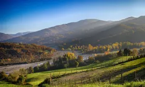 Colline autunnali illuminate dal sole, perfette per il trekking in Emilia Romagna. Un fiume serpeggia tra alberi dai colori caldi.