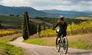 Donna in bici su strada sterrata tra vigneti e cipressi, con colline toscane sullo sfondo. Perfetti giri in bici toscana.