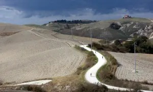 Ciclisti affrontano una strada eroica toscana, serpeggiando tra colline arate sotto un cielo minaccioso.
