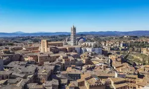 Panoramica di **Siena da vedere**: tetti in cotto, il Duomo e il Campanile svettano su un paesaggio collinare sotto un cielo azzurro.