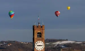 Un colorato evento mongolfiere vola nel cielo azzurro sopra una torre dell'orologio e un paesaggio collinare innevato.