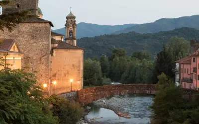 Ponte in pietra sul fiume, con edifici storici illuminati e il monte Argenia sullo sfondo.