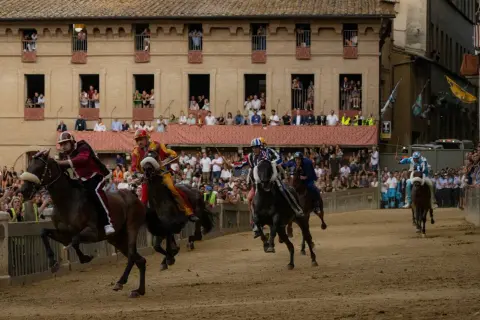 Cavalli e fantini in gara durante le contrade di Siena, con spettatori affacciati ai palazzi.