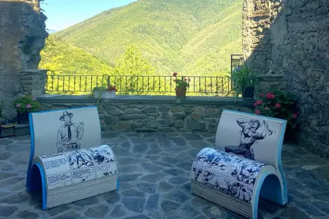 Due sedie a forma di libro con fumetti western, in un cortile con vista sulle colline verdi. Montereggio, paese dei librai, offre un angolo lettura unico.
