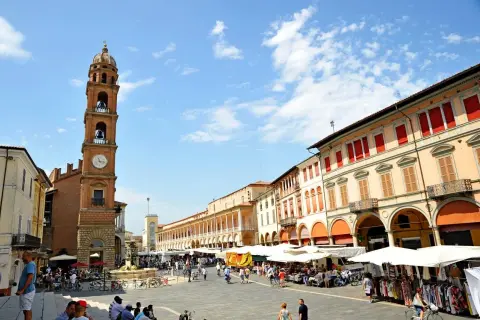Piazza gremita a Faenza, Italia, con un mercato vivace sotto un cielo azzurro. La torre dell'orologio domina la scena.