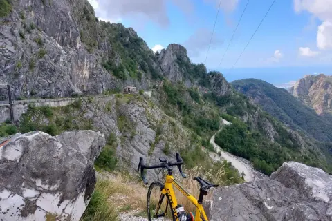 Bicicletta gialla in primo piano, con un panorama mozzafiato di montagne scoscese e un passo del vestito che si snoda tra le rocce.
