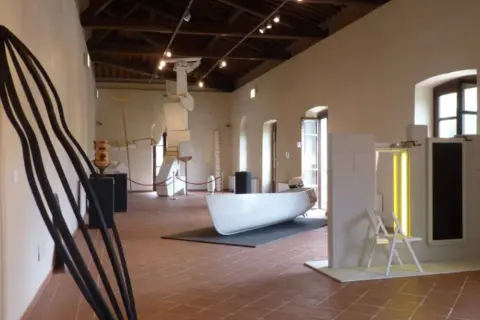 Interno del **mudac carrara** con sculture moderne, tra cui una grande forma nera appoggiata al muro e una barca bianca al centro.
