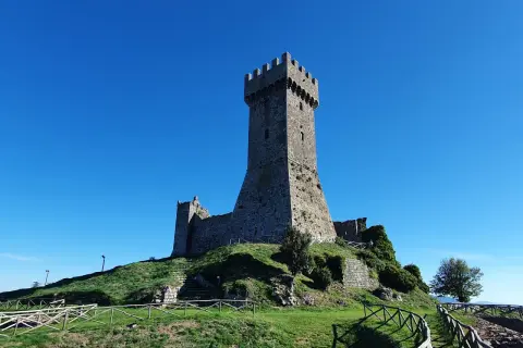 La fortezza di Radicofani domina il paesaggio sotto un cielo azzurro. Le antiche mura e la torre si ergono su una collina verdeggiante.
