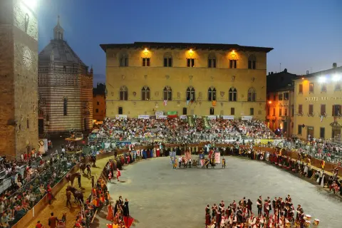 La giostra dell'orso in piazza, con figuranti in costume, cavalli e tribune gremite di spettatori sotto il cielo serale.