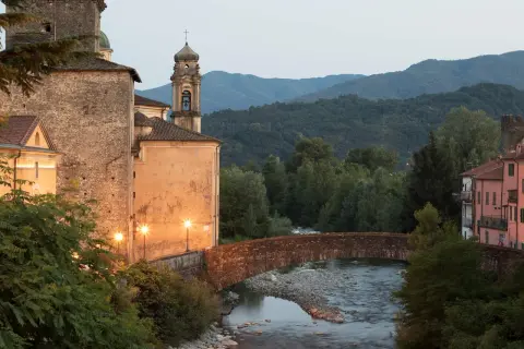 Ponte in pietra sul fiume, con edifici storici illuminati e il monte Argenia sullo sfondo.