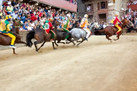 Cavalli e fantini colorati corrono nella polvere del **Palio di Siena**. La folla assiste con passione.