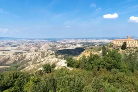 Panoramica delle balze di Volterra, con un antico palazzo in pietra su una collina e dolci colline ondulate all'orizzonte sotto un cielo azzurro.