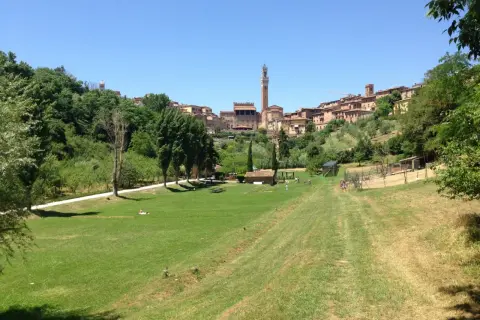Un prato verde con persone che passeggiano, sullo sfondo il belsedere di Siena con i suoi edifici storici e la torre del Mangia.
