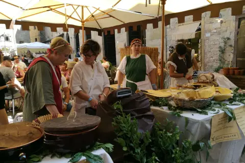 Donne in costume d'epoca servono cibo a un **Volterra markt**. Cesti, pentole e verdure fresche decorano il banco.