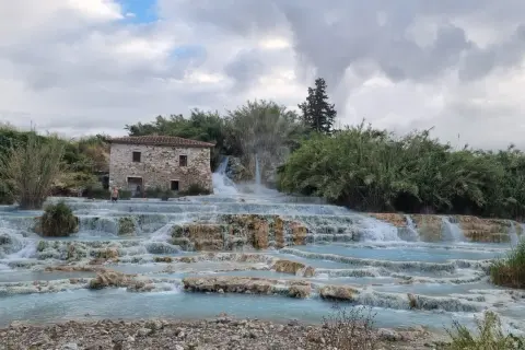 Cascate naturali in Toscana con un antico casale in pietra. L'acqua azzurra scorre su rocce calcaree, creando piscine termali dove alcune persone si rilassano.