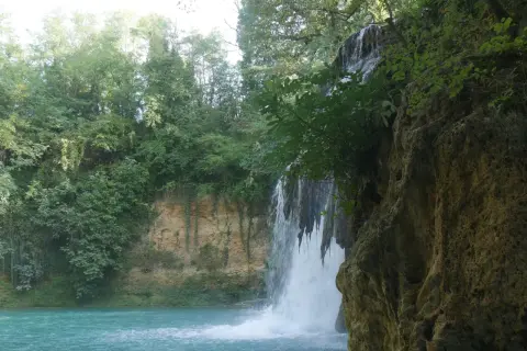 Cascata nel parco fluviale dell'Elsa, con acqua turchese e vegetazione lussureggiante che incornicia la roccia.