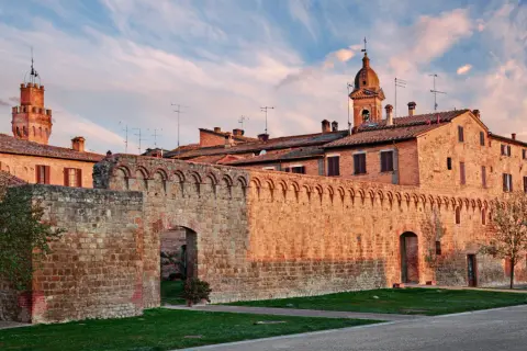 Antica cinta muraria e torri in mattoni rossi sotto un cielo azzurro striato di nuvole rosa, tipico delle colline senesi al tramonto.