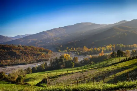 Colline autunnali illuminate dal sole, perfette per il trekking in Emilia Romagna. Un fiume serpeggia tra alberi dai colori caldi.