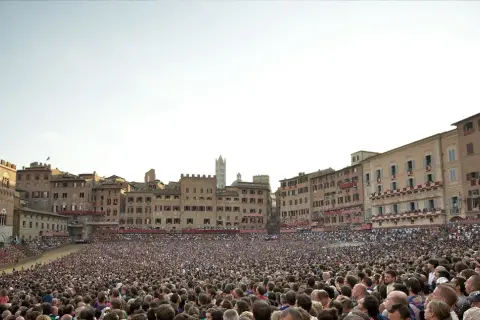 Folla immensa in Piazza del Campo a Siena, in attesa di sapere quand'è il Palio. Edifici storici e cielo terso.