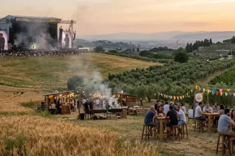Un grande palco per il festival toscana con artisti e folla. In primo piano, bancarelle di cibo e persone che si godono l'atmosfera.