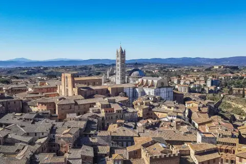 Panoramica di **Siena da vedere**: tetti in cotto, il Duomo e il Campanile svettano su un paesaggio collinare sotto un cielo azzurro.