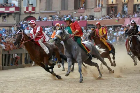Cavalli e fantini in gara durante il Palio di Siena. La polvere si solleva mentre i cavalli corrono verso la vittoria.
