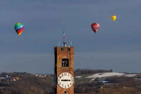 Un colorato evento mongolfiere vola nel cielo azzurro sopra una torre dell'orologio e un paesaggio collinare innevato.