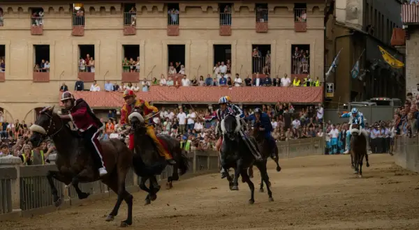 Cavalli e fantini in gara durante le contrade di Siena, con spettatori affacciati ai palazzi.
