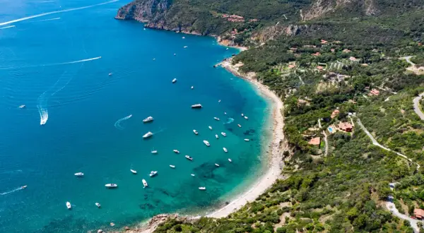 Baia con acque cristalline e numerose barche al largo della costa del Monte Argentario. Ville immerse nel verde e una strada panoramica.