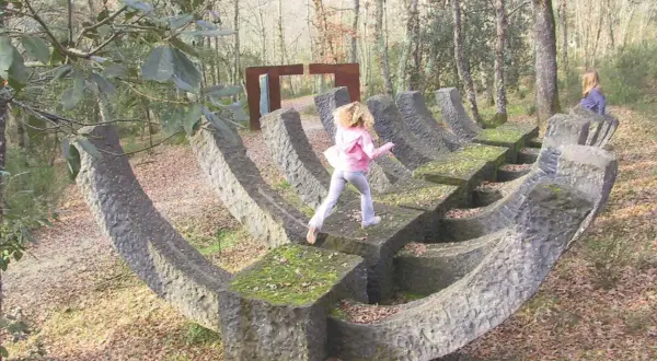 Bambini esplorano un **museo a cielo aperto in Toscana**, tra sculture in pietra e alberi secolari.