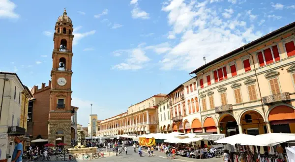 Piazza gremita a Faenza, Italia, con un mercato vivace sotto un cielo azzurro. La torre dell'orologio domina la scena.