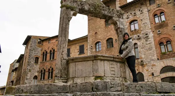 Donna sorride vicino a un antico pozzo a San Gimignano, con palazzi medievali sullo sfondo.