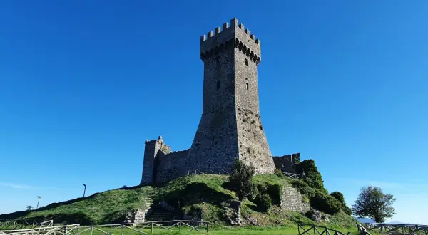 La fortezza di Radicofani domina il paesaggio sotto un cielo azzurro. Le antiche mura e la torre si ergono su una collina verdeggiante.