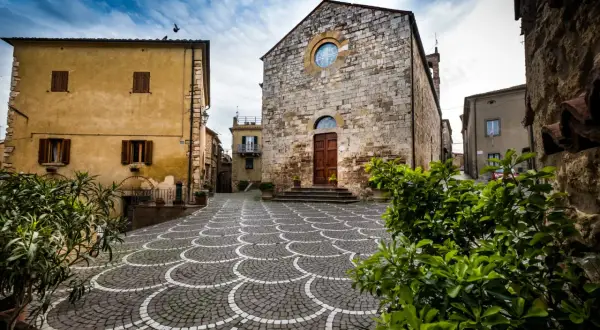 Piazza con pavimentazione a ventaglio, chiesa in pietra e case gialle, tipici borghi vicino Marina di Bibbona.