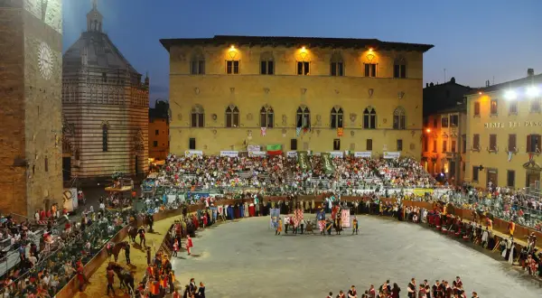 La giostra dell'orso in piazza, con figuranti in costume, cavalli e tribune gremite di spettatori sotto il cielo serale.