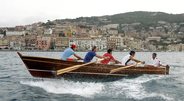 Equipaggio in gara nel palio marinaro argentario, vogando all'unisono con la città sullo sfondo.